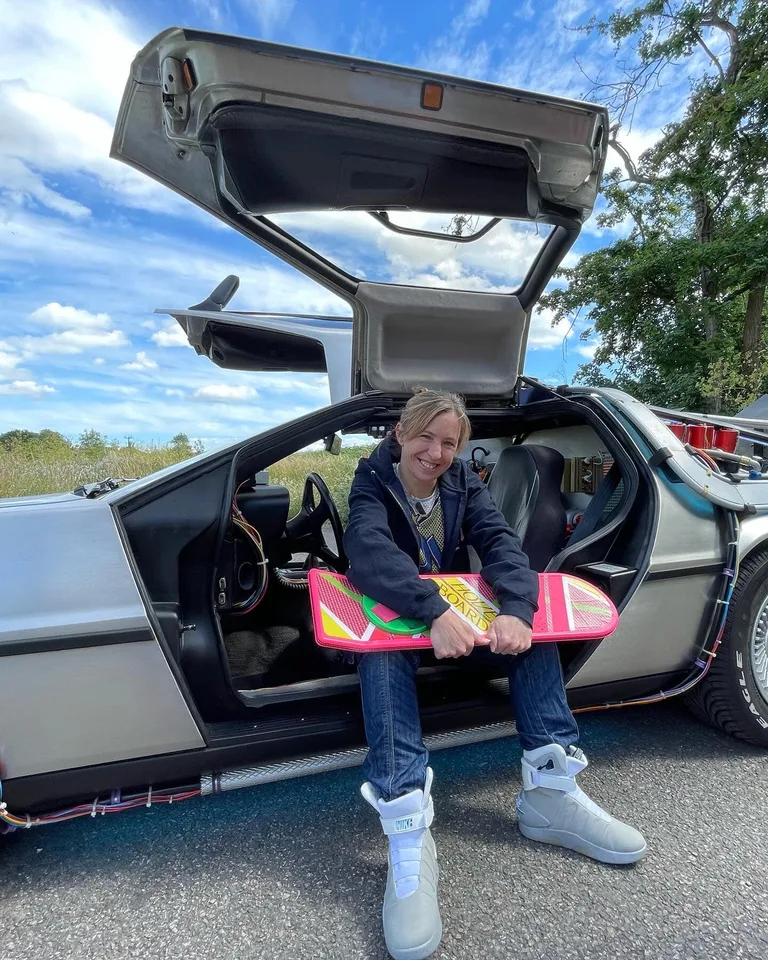 Marty McFly tribute pose — guest with hoverboard sitting on the open DeLorean gull-wing — rent for 80s-themed birthdays