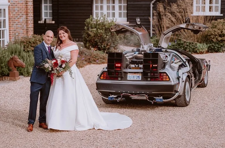 Newly-weds standing beside the DeLorean Time Machine with gull-wing doors up and OUTATIME plate — wedding car hire
